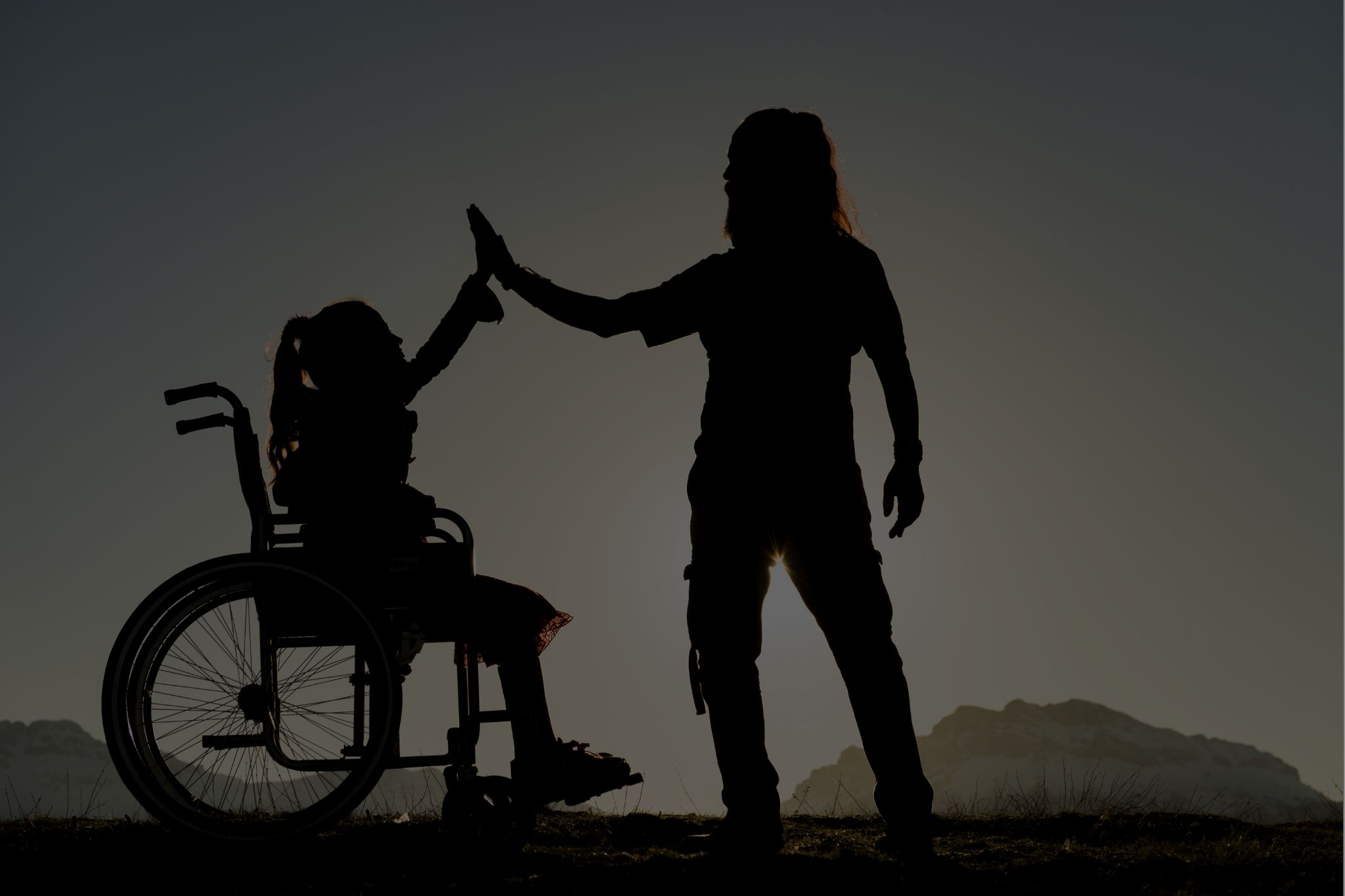 Silhouette of a girl in a wheelchair high-fiving an adult at sunset